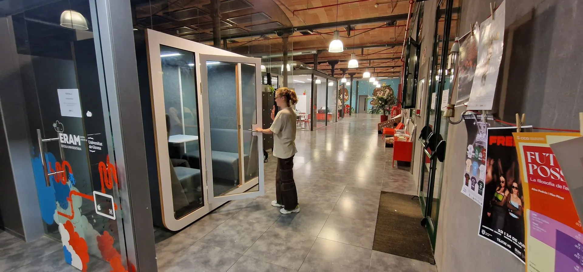 A person opening the glass door of a white Silentbox Duet booth in a coworking space with an exposed brick ceiling.